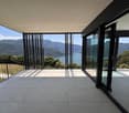 Large balcony or terrace with panoramic views of a mountainous landscape and a body of water, such as a lake or ocean. The space features floor-to-ceiling glass windows and doors, providing an open and unobstructed view of the scenic surroundings.