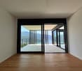 Spacious living room with large windows and sliding glass doors leading to a balcony or terrace, offering a scenic view of the surrounding mountains or landscape. The room has hardwood flooring and a minimalist, modern design.