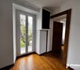 This image shows a room with a large window overlooking a balcony or terrace. The room has white walls and hardwood floors. There is a wooden door frame visible in the background.
