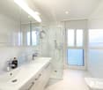 Spacious bathroom with white tiles, large vanity sink, glass shower enclosure, and large windows providing natural light