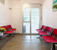 The image shows a waiting room or lobby area with red leather chairs arranged in rows. There is a large window with a view of the outdoors, and a small table with a vase of flowers. The floor appears to be a concrete or tile material.
