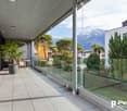 Spacious balcony with glass railing, overlooking a landscaped garden and snow-capped mountains in the background. The apartment features a modern, minimalist design with large windows allowing for abundant natural light and scenic views.