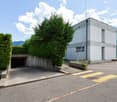 A modern, two-story commercial or industrial building with a garage entrance, surrounded by landscaping including tall hedges and trees.