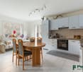 The image shows an open-plan kitchen and dining area. The kitchen features white cabinets, a tiled backsplash, and a built-in oven and stovetop. The dining area has a round wooden table with four chairs. The room has large windows that provide natural lig
