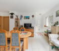 Open-concept living room and dining area, with wooden furniture including a dining table and chairs, a TV stand, and shelving. The room has white walls, tile flooring, and a sliding glass door leading to a balcony or terrace.