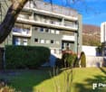 4 story building, green exterior, multiple balconies, brick wall, small house on the side, garden in the front, lawn, bushes