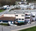 The image shows a commercial/industrial area with several buildings and structures. There is a road running through the area with parked vehicles visible. The buildings appear to be a mix of commercial and industrial structures, with some having a more mo