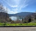 The image shows a scenic view of a lake surrounded by mountains and forests. The lake appears to be a vibrant turquoise color, contrasting with the bare trees and lush green vegetation on the slopes. The sky is partly cloudy, adding to the picturesque nat