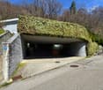 A unique, partially underground building with a curved, ivy-covered roof. The building has a stone exterior and a paved entrance area with benches. The surrounding area is forested with bare trees, indicating a spring or fall season.