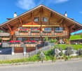 2 story wooden house, red flowers on balconies, fence in front, green grass, clear sky