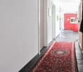 narrow hallway with white walls, tile flooring, red rug, open doorways, a red kitchen