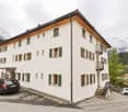 3-story apartment building with white exterior, green shutters, and a slanted roof. There is a parking area in front of the building with a few cars parked. The building is situated in a mountainous, forested area with snow-capped peaks visible in the bac