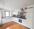 Spacious L-shaped kitchen with white cabinets, granite countertops, stainless steel appliances including a dishwasher, and a large window providing natural light. The kitchen has a hardwood floor and an open layout.