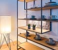 A shelf unit in a room with various plates, cups, wine glasses, and small potted plants. A floor lamp on the left side of the shelf and an electrical outlet at the bottom of the shelf.