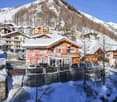 Snowy mountain area with buildings, a car park, a small stream, and mountains in the background.