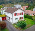 Aerial view of a white house with brown roof, red shutters, a small attached garage and greenery surrounding it.