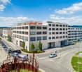 Large office building with multiple floors, modern architecture, glass windows, and a rooftop terrace. Located in an urban area with a busy road and parking lot.