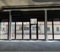 The image shows a large commercial space with floor-to-ceiling windows overlooking a city street. The space appears to be unfinished, with exposed concrete walls and ceiling. The windows provide a clear view of the surrounding buildings.