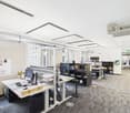Modern office setup with white desks, adjustable height tables, various office equipment and chairs, large windows