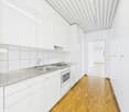 Bright and modern kitchen with white cabinets, stainless steel appliances including a dishwasher, and a tiled backsplash. The kitchen leads to a hallway with white walls and a wooden floor.