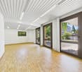 Large open space with high ceilings, wooden flooring, and large windows overlooking a garden area. The room has a modern and minimalist design with a clean, bright atmosphere.