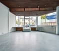 Empty room with tiled floor, white walls, wooden ceiling, glass doors, and large windows with a view of the street outside.
