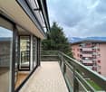 The image shows a balcony or terrace of an apartment building. The balcony has glass sliding doors and a railing. The building has a modern design with a mix of light and dark colors. There are other apartment buildings visible in the background, surround