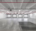 Empty parking lot with white walls, ceiling with pipes, 4 garages, emergency door on right, square gray mat
