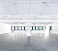 Large open space with white walls, gray hardwood floors, and multiple windows. The room has a minimalist design with no furniture or decor visible.