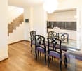 glass dining table, glass chairs, wooden floor, white walls, kitchen with white cabinets, stainless steel sink, staircase with glass railing