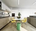 The image shows a modern, open-concept kitchen and dining area. The kitchen features white cabinets, a marble backsplash, and a black oven. The dining area has a white table with yellow chairs, and a green stool. There is also a potted plant and some artw