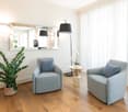 Two armchairs, a table, a plant, a mirror, a lamp, wooden floor, white walls, white curtains