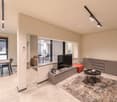 Open concept living space with living room, dining area, and kitchen. Hardwood floors, modern furniture, and large windows providing natural light. TV console, coffee table, and side tables in the living room area. Dining table with chairs. Sliding glass