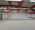 Large open commercial space with high ceilings, exposed red metal beams and supports, and a gray carpeted floor. The space appears to be a warehouse or industrial-style office area with ample natural lighting from the windows.