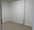 Empty room with white walls and carpeted floor. White ceiling with a single fluorescent light. Electric outlet mounted on the wall.