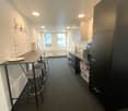 The image shows a studio apartment with an open floor plan. The kitchen area features a black refrigerator, a dishwasher, and a counter with bar stools. The living area has a table and chairs. The apartment has white walls, recessed lighting, and dark gra