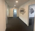 The image shows a hallway or corridor in an office space. The walls are painted in a light color and have a large graphic design with words related to teamwork, collaboration, and productivity. The floor is covered in dark gray carpeting. The hallway lead