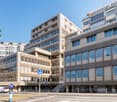 A large office building with multiple floors and many windows, some with white doors, situated on a raised ground floor
