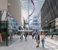 The image shows a modern commercial building with a glass facade and a covered walkway. Several people are walking through the walkway, which is lined with shops and businesses. The building has a sleek, contemporary design with a mix of glass and metal m