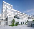 Multi-story office building with a modern, minimalist design featuring large windows, white exterior, and a landscaped courtyard with trees and greenery. The building appears to have a parking area or garage visible in the foreground.
