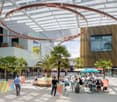 Large indoor commercial space with glass walls, palm trees, outdoor seating area, and a circular overhead lighting structure