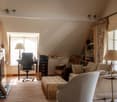 Cozy living room with large windows, beige sofa, armchair, coffee table, desk with chair, bookshelves, and curtains. The room has a warm, inviting atmosphere.