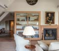 Cozy living room with a fireplace, built-in wooden bookshelves, a white armchair, and a framed painting on the wall. The room also features a dining area with a wooden table and chairs.