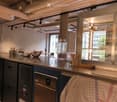 modern kitchen with stainless steel countertops, various kitchen utensils, cabinet drawers, a dishwasher, a large glass door with shelves, and a hanging rack.
