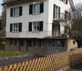 Two story house, brown roof, balcony, fenced garden, garages, white exterior, green windows, chimney, grass lawn, wooden fence