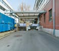An industrial building with a covered loading bay, surrounded by brick walls and a concrete path, containing blue dumpsters, steel structures, and pallets.