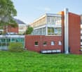 Multi-story red brick building, large windows, modern architecture, surrounded by grass and trees, HVAC units on the side