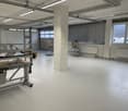 Large open industrial space with concrete floors, metal shelving units, and large windows providing natural light. The space appears to be used for storage or manufacturing purposes.