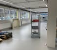 The image shows an open office space with a concrete ceiling, fluorescent lighting, and large windows. There is a metal storage rack and some other equipment visible in the room.