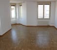 Empty room with wooden floors, white walls, three windows, and radiators.